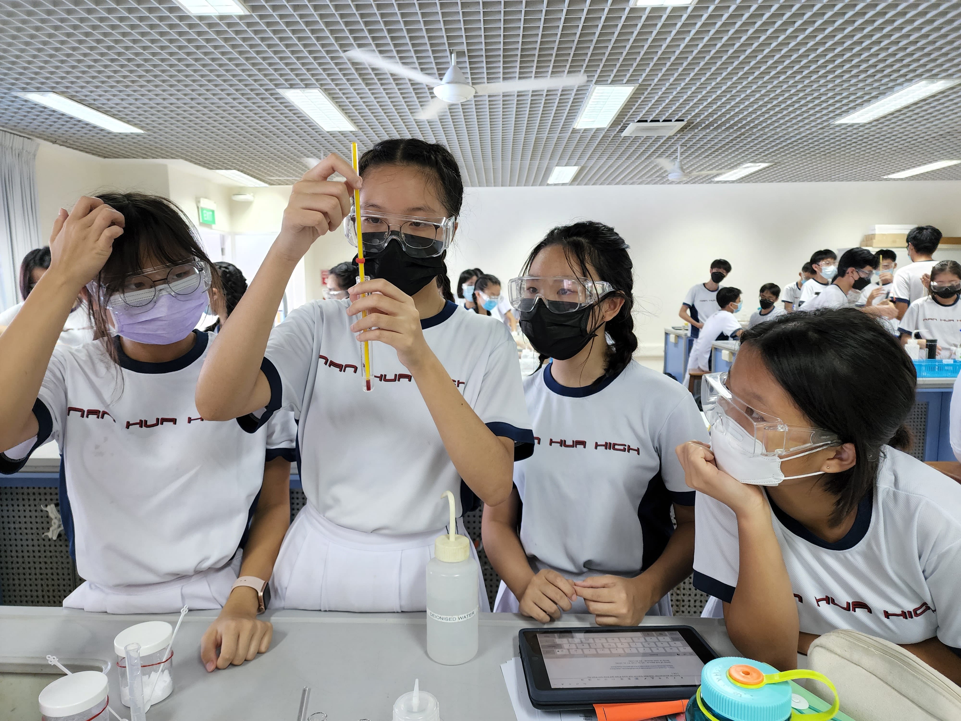 Students in Nan Hua High uniforms wearing goggles and masks conduct a science experiment with a thermometer in a lab.